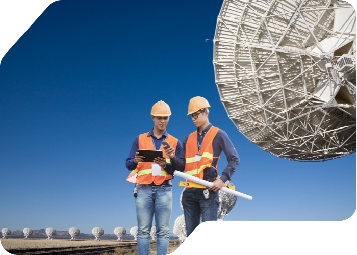 Two men in hard hats stand in front of a large satellite dish, discussing its installation and maintenance
